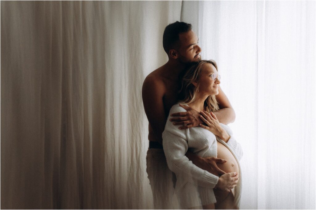 Couple enlacé en peau à peau devant une fenêtre lors de leur séance photo maternité à domicile.