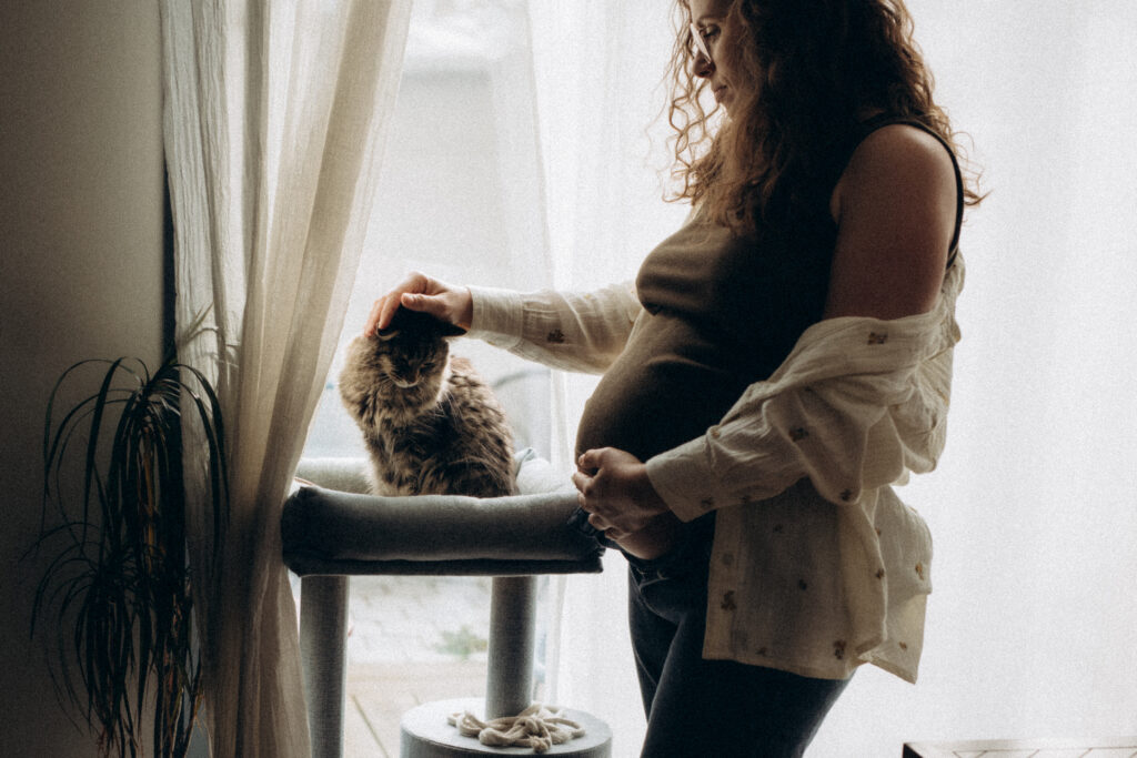 Une femme enceinte caresse son chat devant une fenêtre.