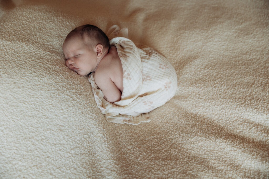 Un bébé allongé sur un plaid beige. Il est sur le ventre endormi et emmailloté.