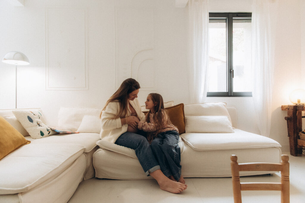 Une maman et sa fille souriant et assises sur un canapé beige.