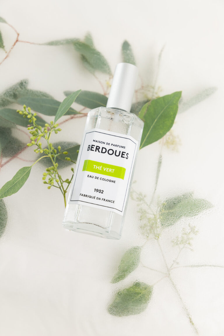 Un parfum transparent posé sur une vitre humide et sur un ensemble de feuilles vertes.