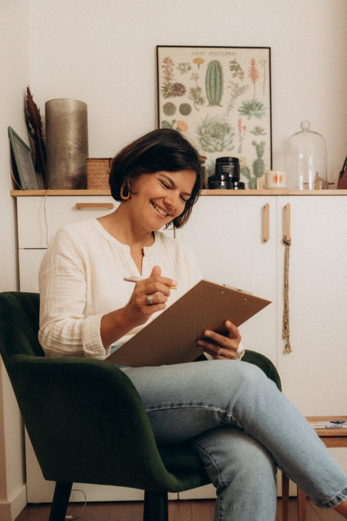 Une femme assise sur une chaise noire et écrivant sur un carnet beige.