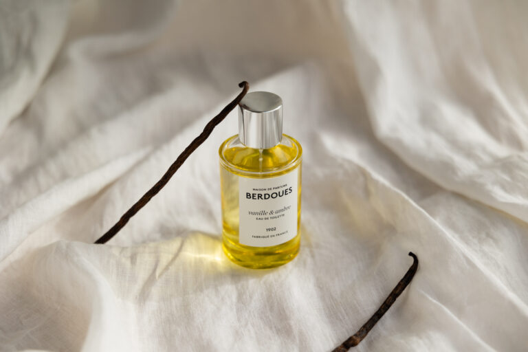 Un parfum jaune posé sur un tissu beige plissé et entouré de gousses de vanille.