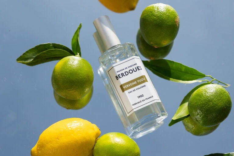 Un parfum posé sur un miroir dans lequel se reflète le ciel et entouré de citrons verts et jaunes.