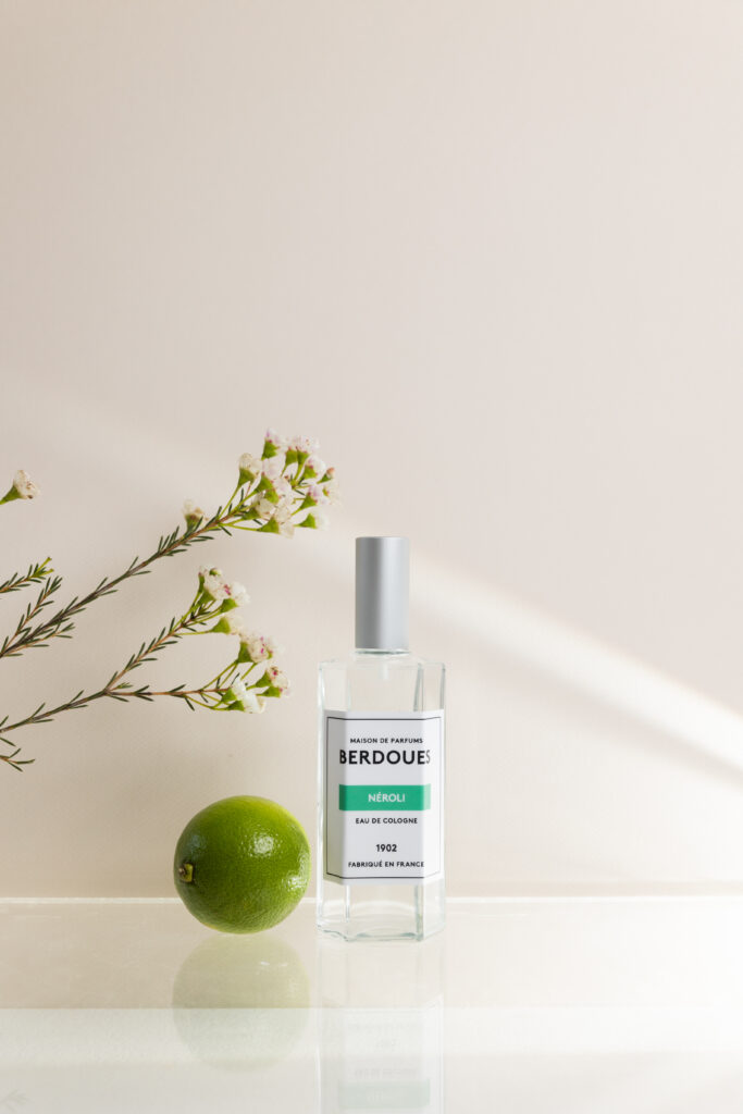 Un parfum posé sur une vitre avec un faisceau lumineux qui le traverse. Il est entouré d'une fleur et d'un citron vert.