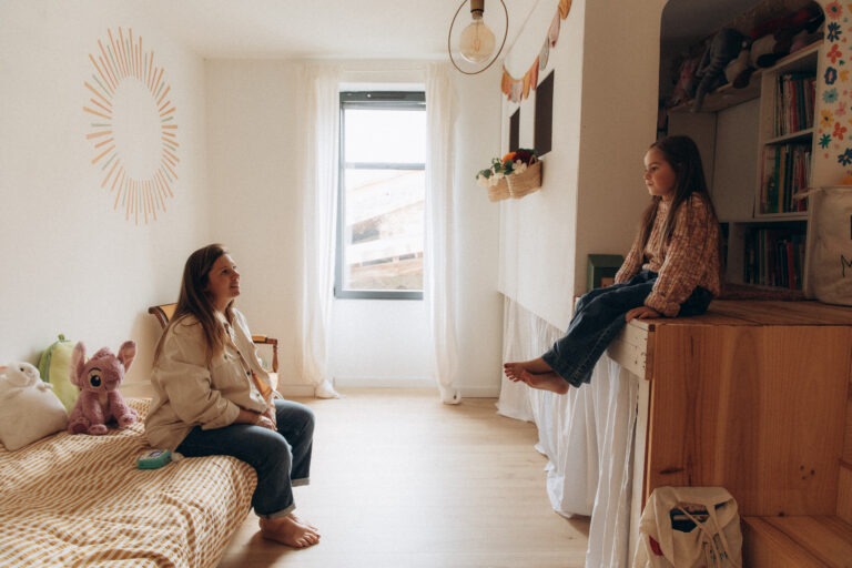 Une maman assise sur le lit d'une chambre d'enfant regarde sa fille assise en face d'elle.