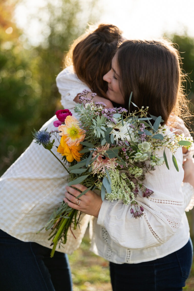 Deux femmes portant des blouses blanches s'enlacent au coucher du soleil avec un bouquet de fleurs à la main.