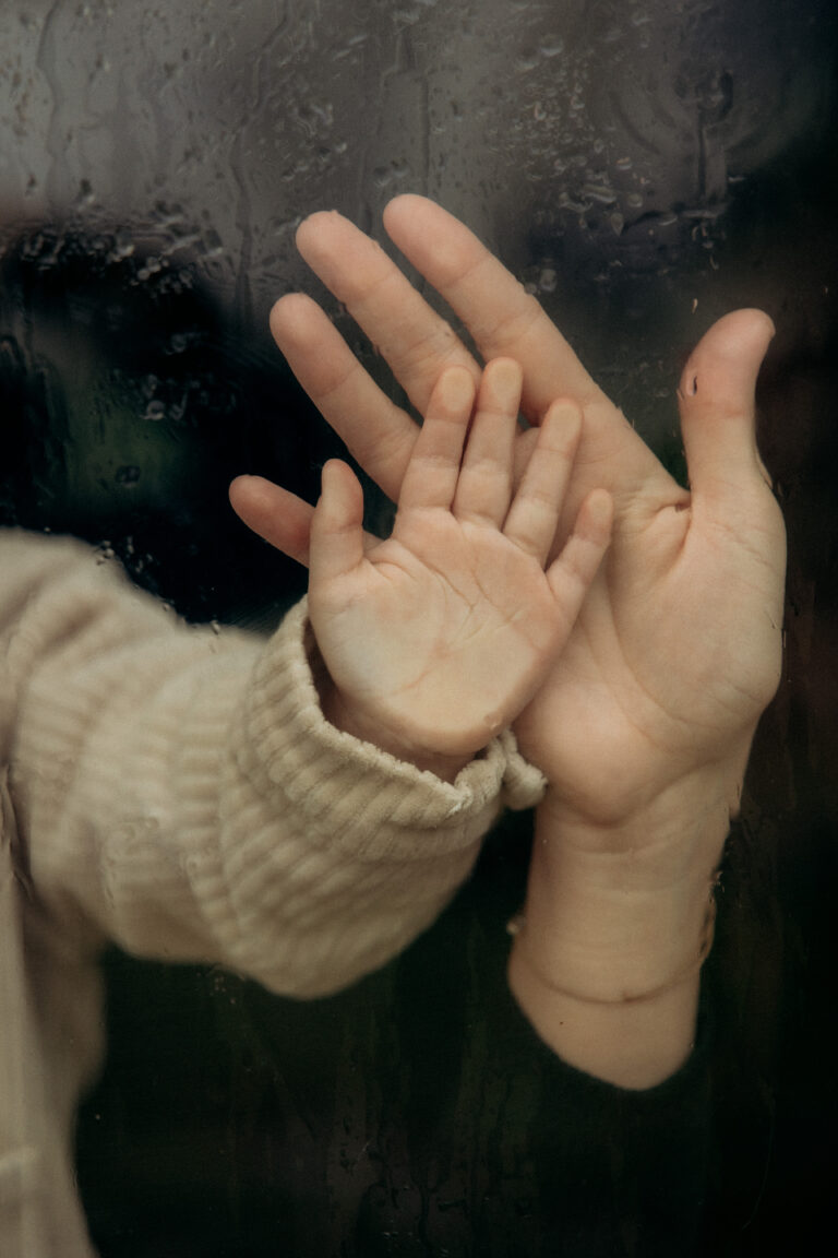 Une main de maman posée sur la main d'un enfant posé sur une vitre mouillée.